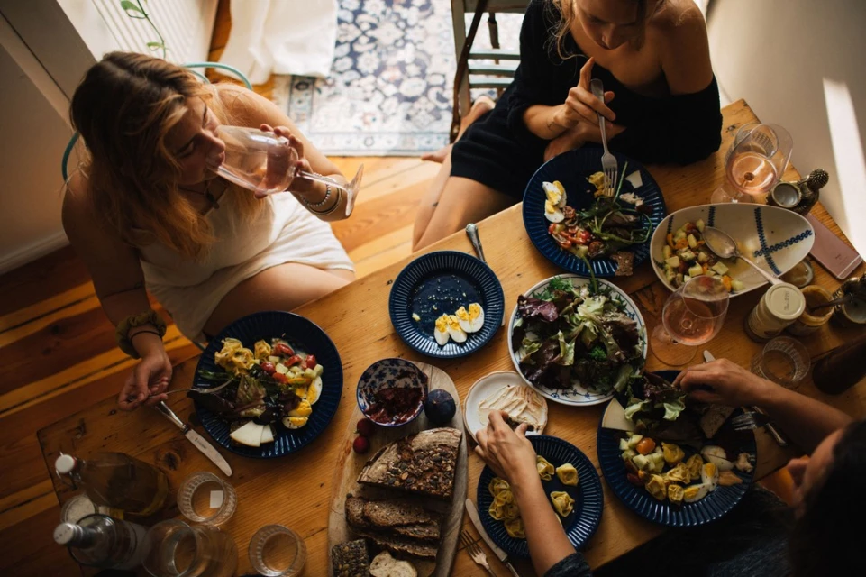 Overhead shot of four people around a wooden table, eating salads, bread, and pasta with drinks in a naturally lit indoor setting.