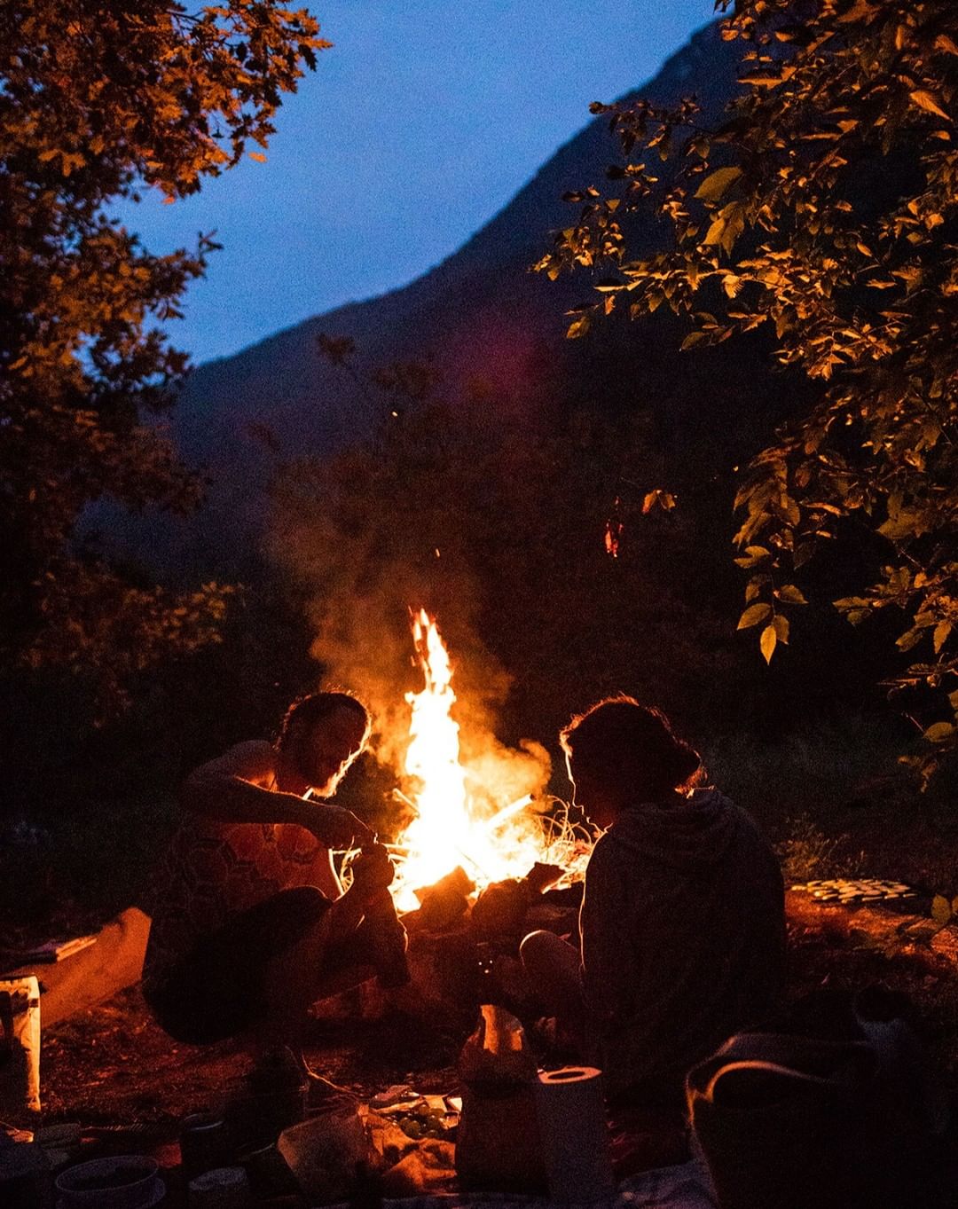 Two people sit near a blazing campfire at night in a wooded area, with a mountain visible in the dark twilight sky.