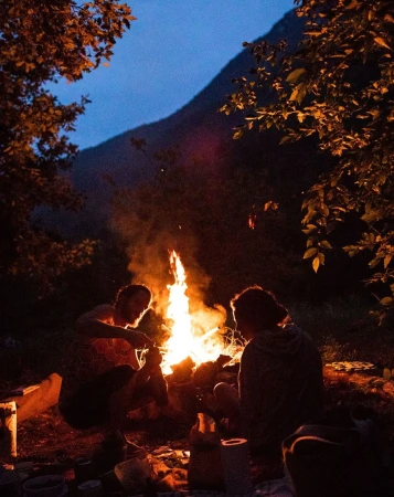 Two people sit near a blazing campfire at night in a wooded area, with a mountain visible in the dark twilight sky.