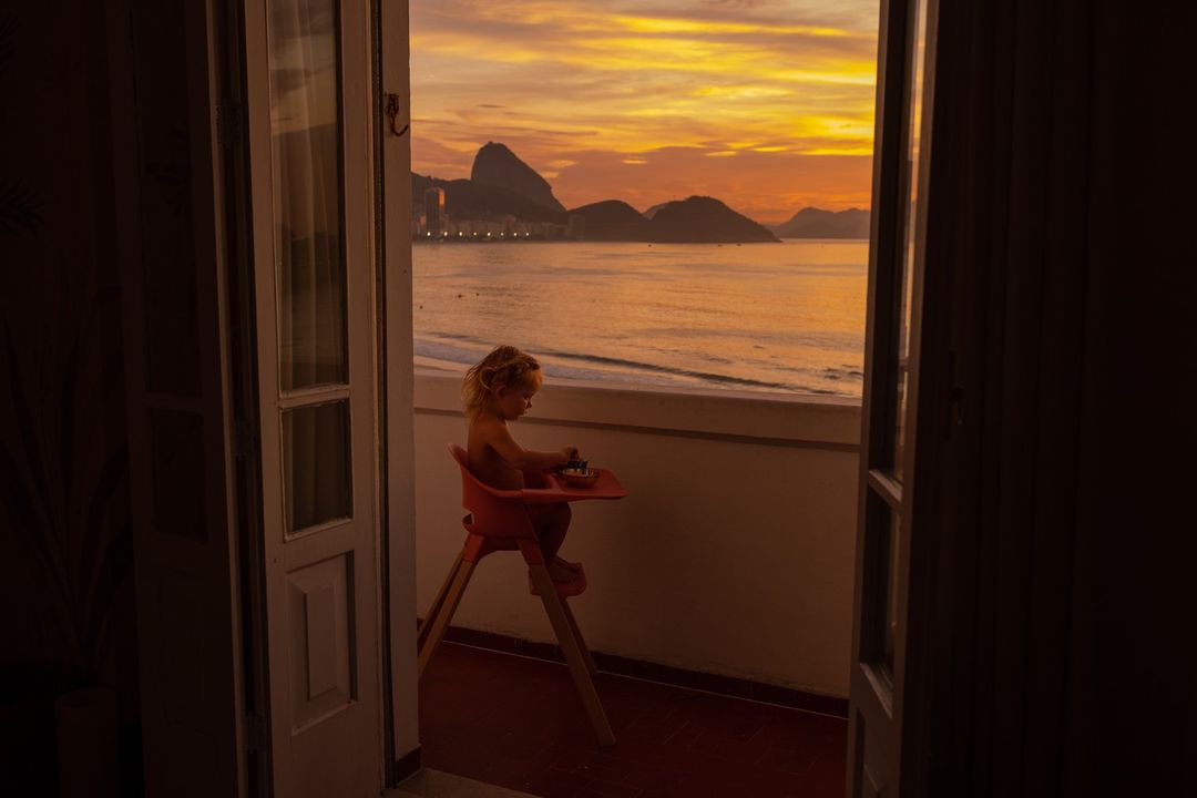A child sits in a high chair on a balcony overlooking the ocean and the silhouetted mountains under a vibrant orange and yellow sunset sky.