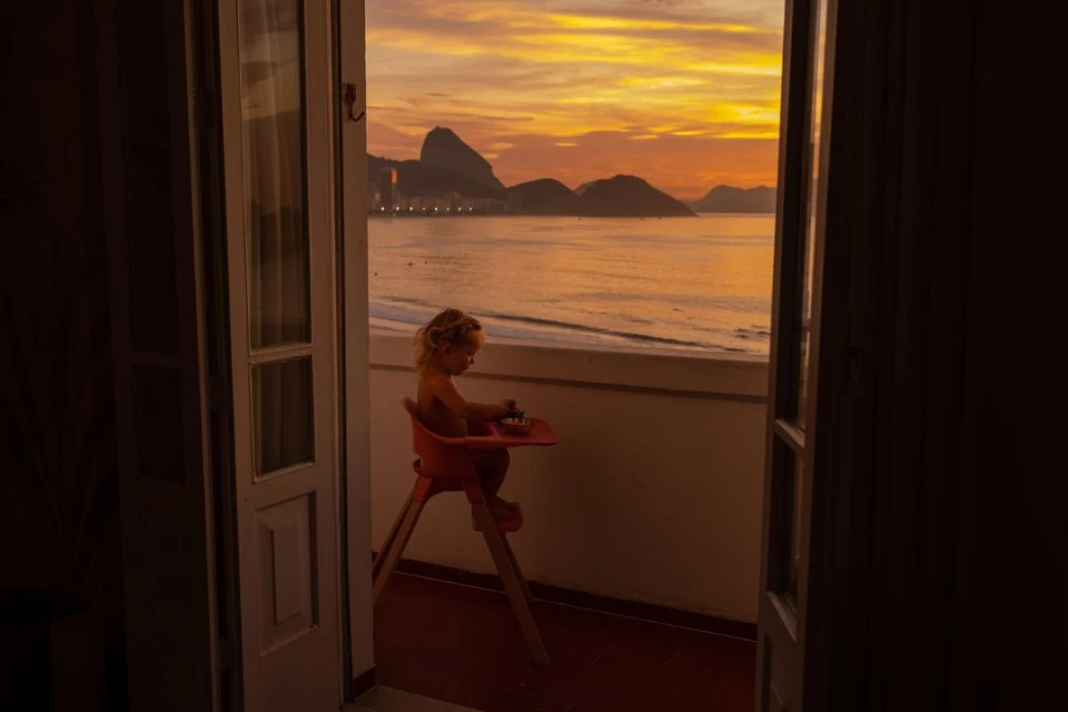 A child sits in a high chair on a balcony overlooking the ocean and the silhouetted mountains under a vibrant orange and yellow sunset sky.
