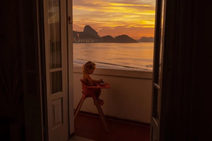 A child sits in a high chair on a balcony overlooking the ocean and the silhouetted mountains under a vibrant orange and yellow sunset sky.