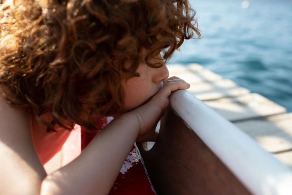 A close-up of a child with curly red hair leaning against a white rail, looking out at a wavy body of water on a sunny day.