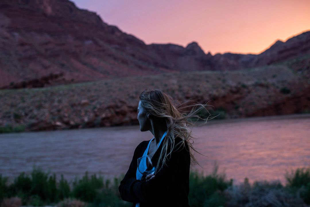 A young woman with long hair stands with her arms crossed, looking out at a river surrounded by canyons during a colorful sunset.