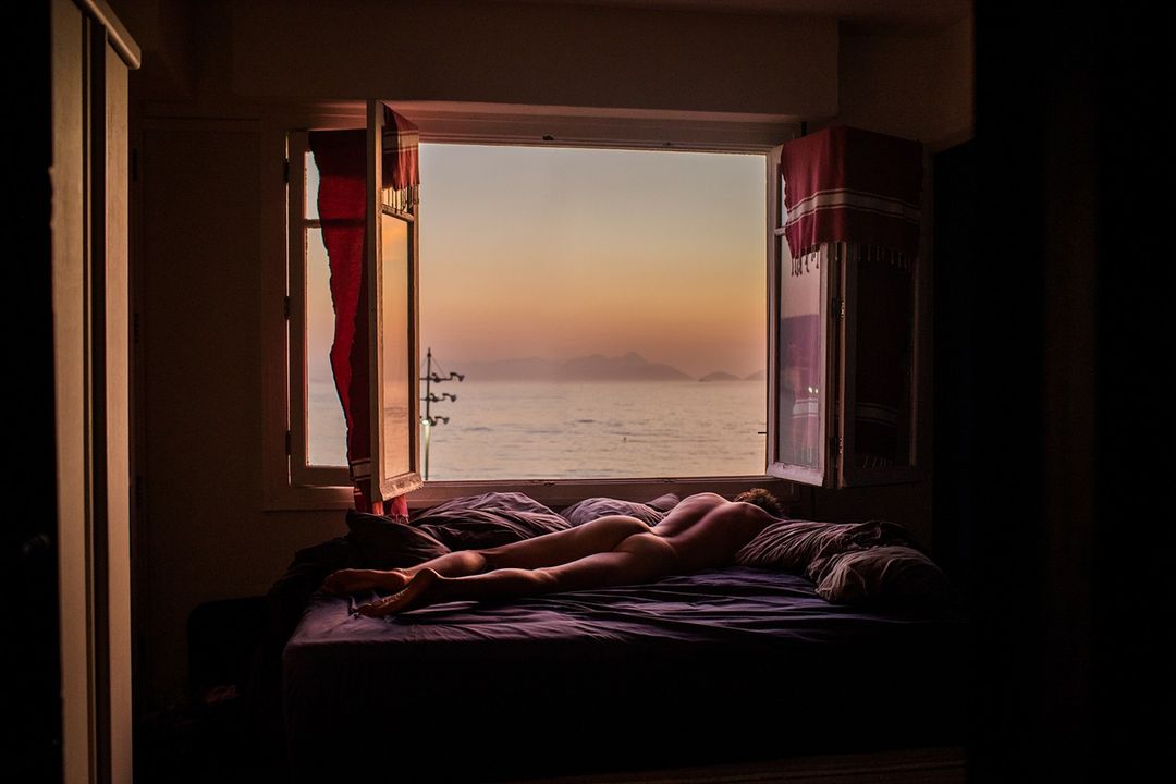 Nude adult lying on bed in bedroom, illuminated by window light with ocean view at sunset, creating a warm and serene artistic photograph.