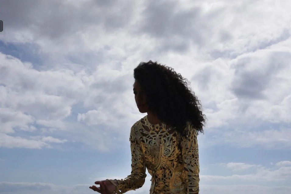 Adult woman with curly hair in ornate gold dress stands outside against a cloudy sky in a fashion or beauty shot.