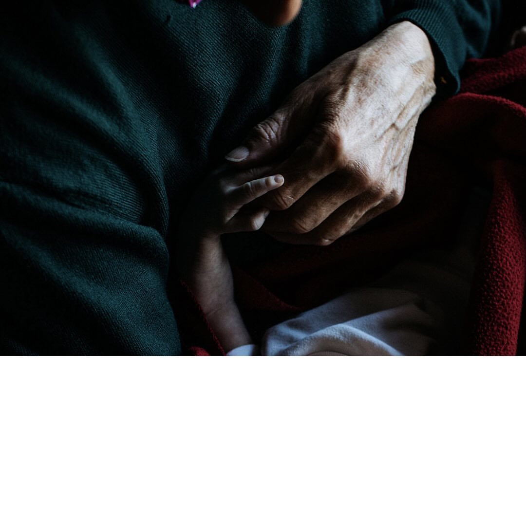 Close-up photo showing an elderly adult hand holding a child's hand, on a lap with red and white fabric; conveying a sense of tenderness.