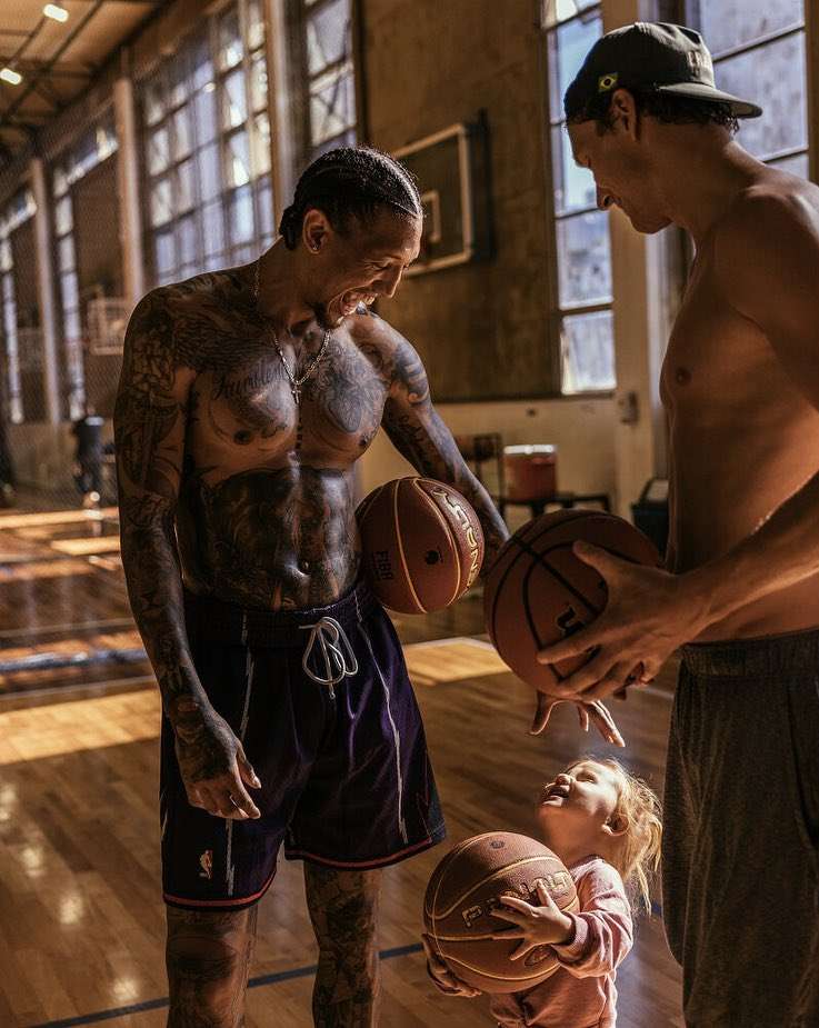 Two tattooed adult men with a child holding basketballs indoors, inside a gymnasium with windows, and wooden floor.