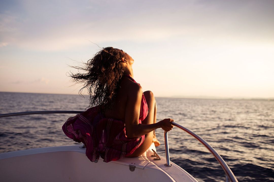Woman with curly hair sits on a boat enjoying the sunset. Wearing a pink wrap, she gazes upward at the orange and pink sky.