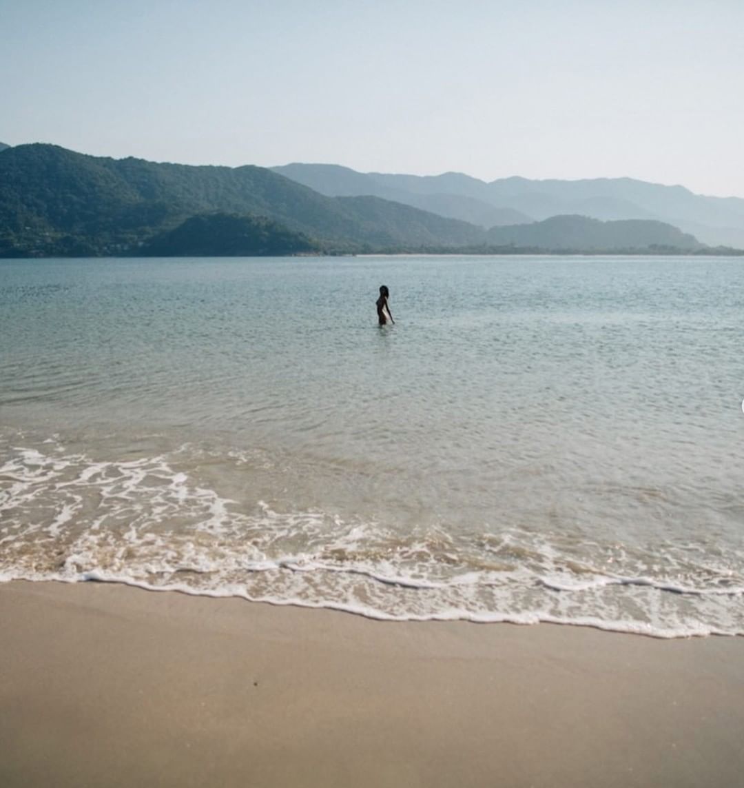 A lone figure wading in calm coastal shallows before distant mountain ridges under an overcast sky.