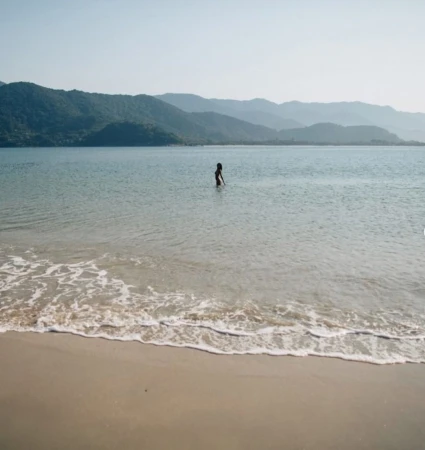 A lone figure wading in calm coastal shallows before distant mountain ridges under an overcast sky.