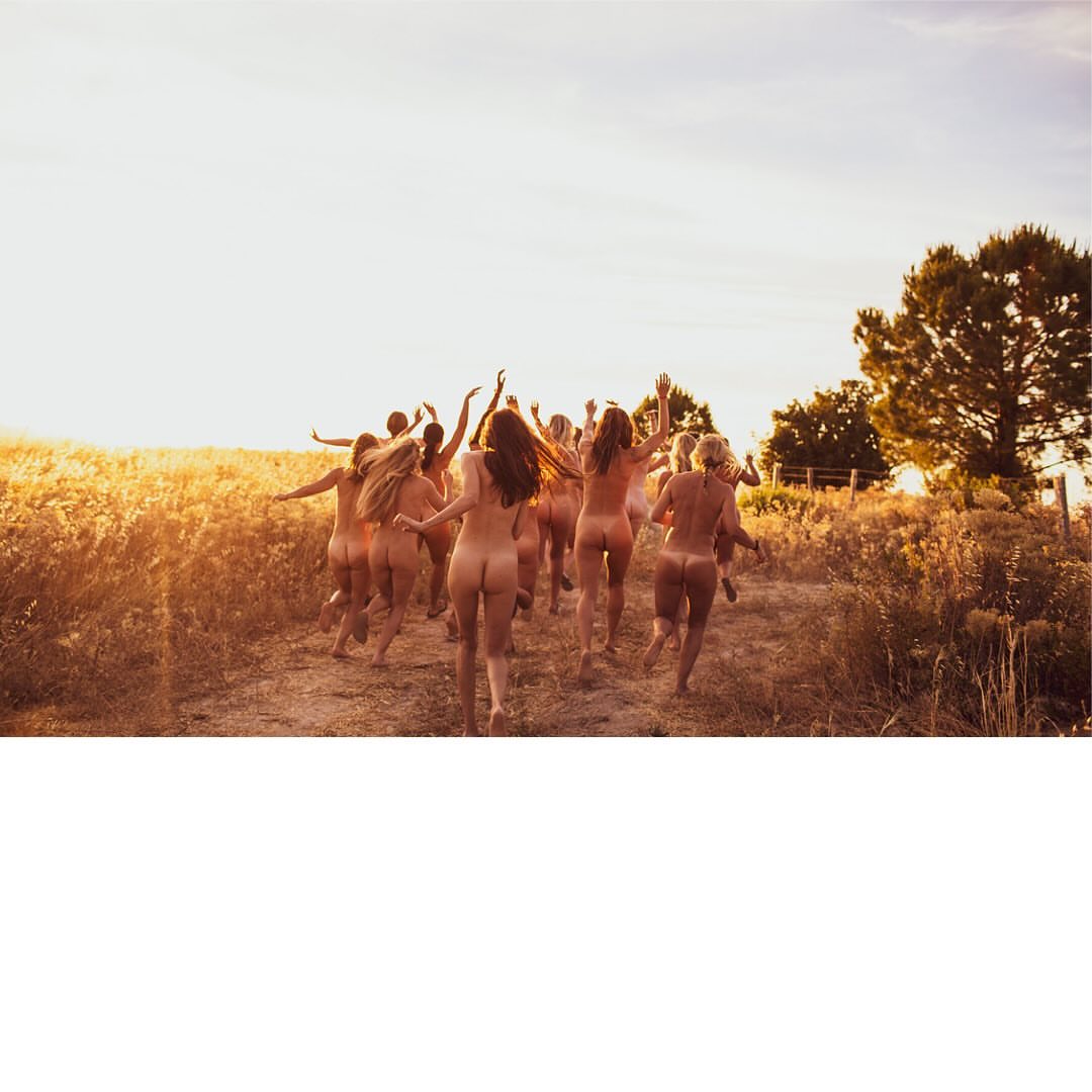 Group of nude adult females running through tall grass in a golden field under a bright sky at sunset, outdoors in a rural setting.