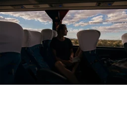 Man seated on a bus looking out the window at a cloudy sky and landscape