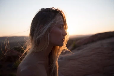 Profile portrait of a blonde woman in silhouette against a bright sunset sky and rocky terrain background.