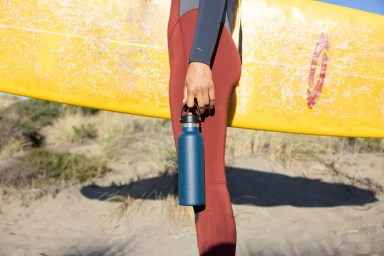 Person holding a blue water bottle wearing a red wetsuit while carrying a yellow surfboard on a sandy beach.