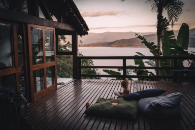 A young child reading a book on a wooden deck of a cabin overlooking a calm body of water during sunset.