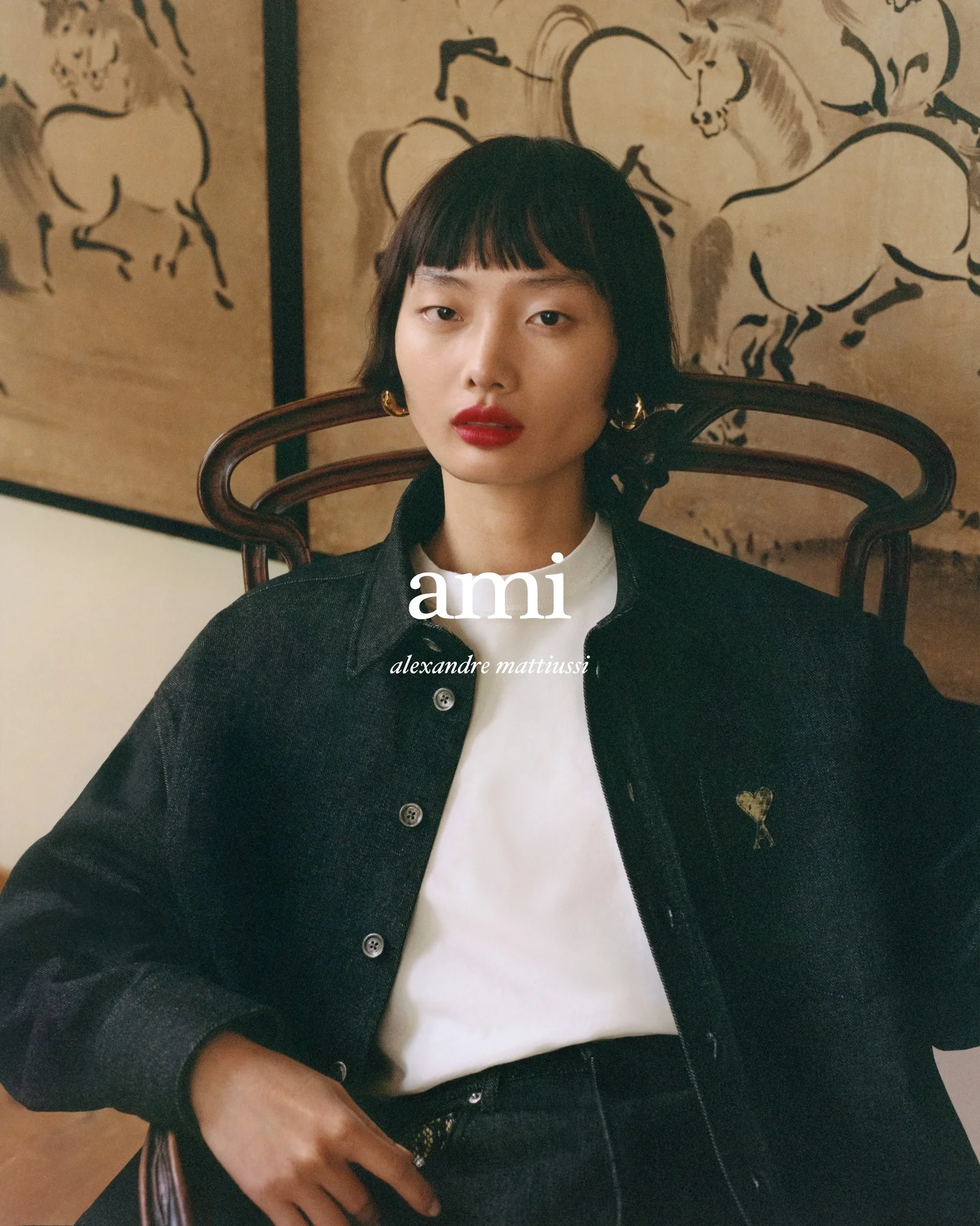 An Asian woman with red lipstick poses indoors wearing a dark denim jacket with a white t-shirt and a brown patterned background.