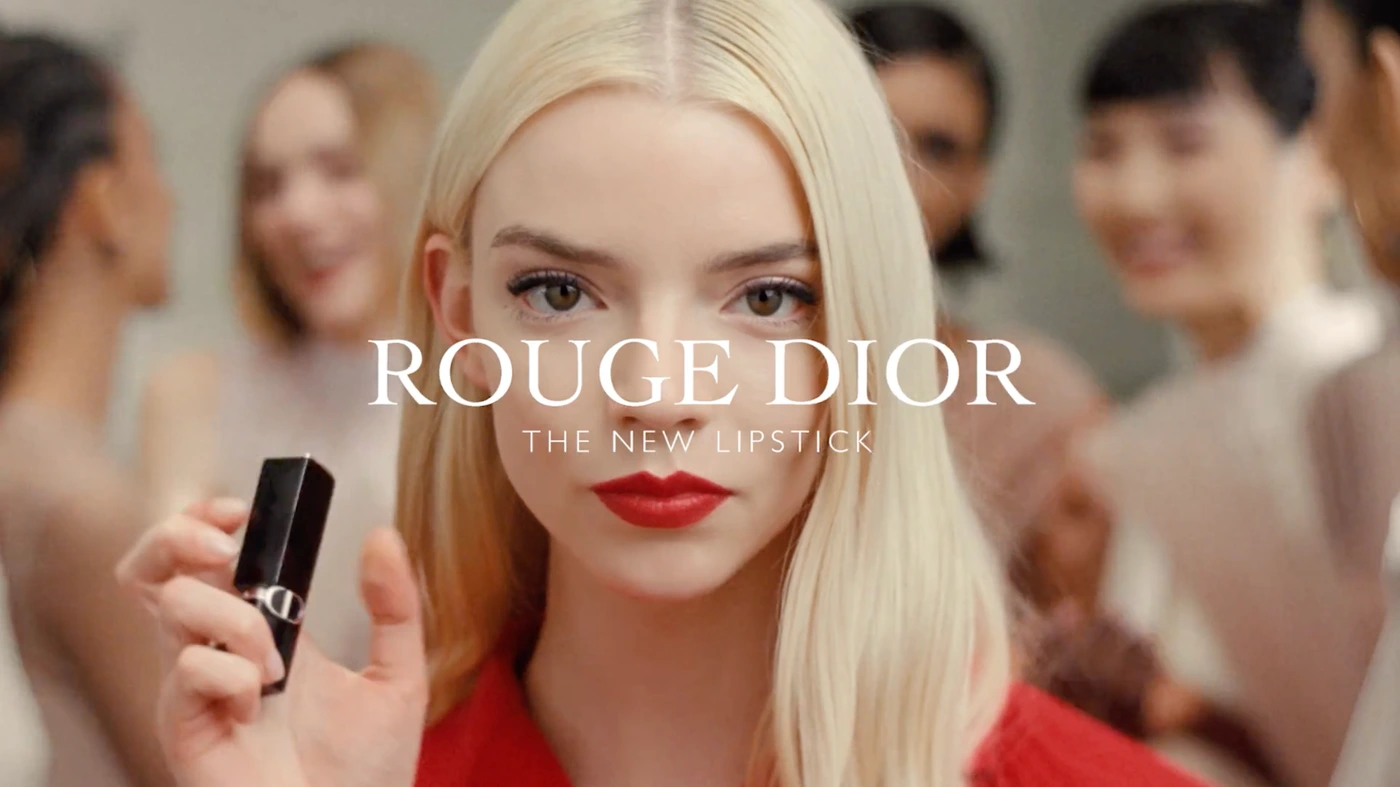 A blonde woman wearing a red top holding a black lipstick tube against a soft-focus background with other people.