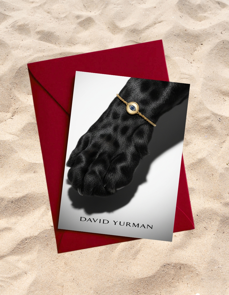 Gold bracelet with blue gemstone on a black dog's paw presented on a greeting card next to a red envelope against a sand background.