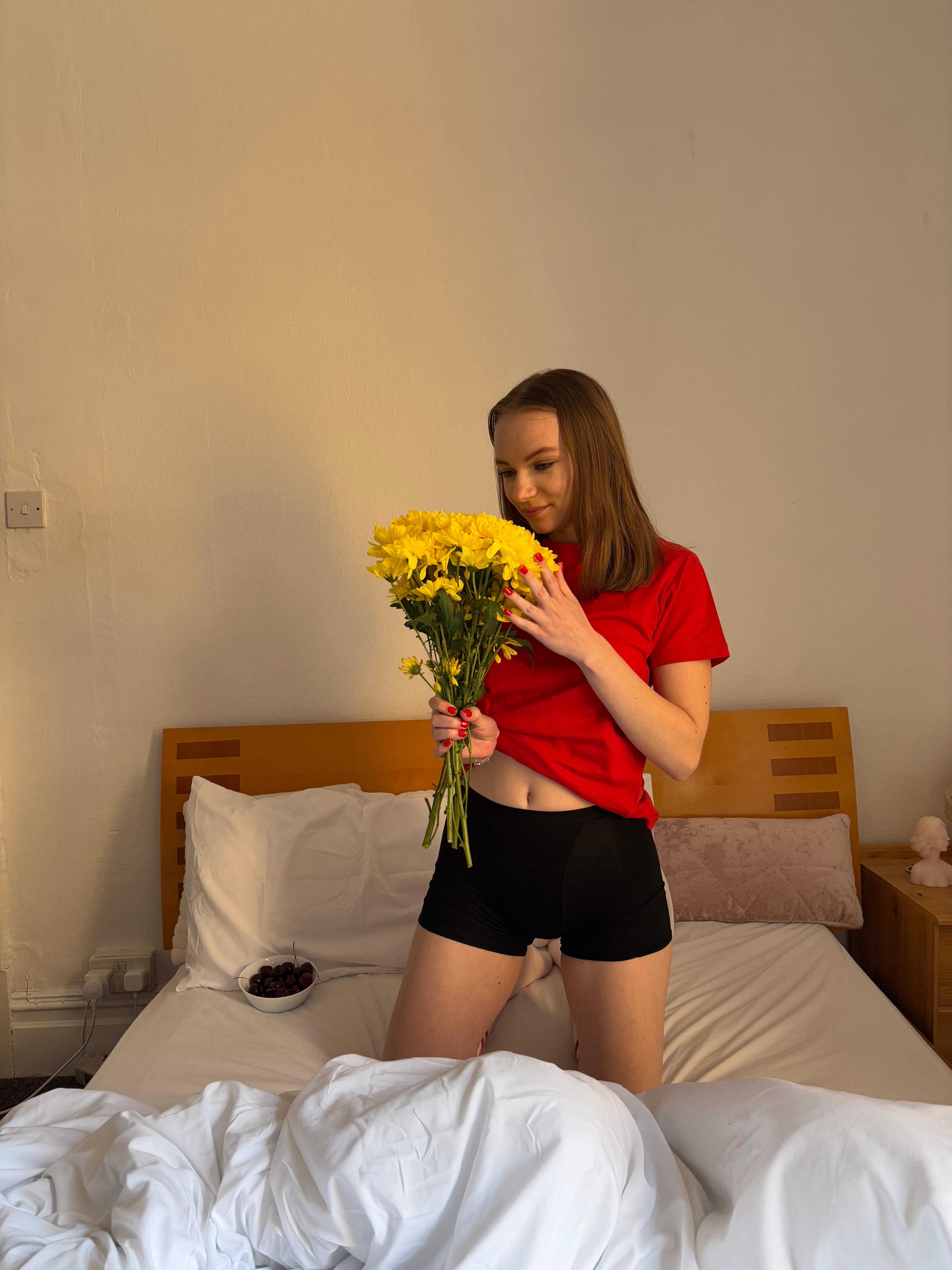Young woman in red shirt holds a bouquet of yellow flowers while kneeling on a bed with white sheets in an indoor setting.