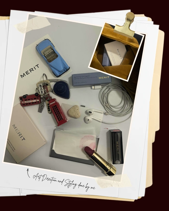 Flat lay composition featuring Merit makeup, a dark lipstick, vintage toy car, London keychains, and wired earphones on white paper.