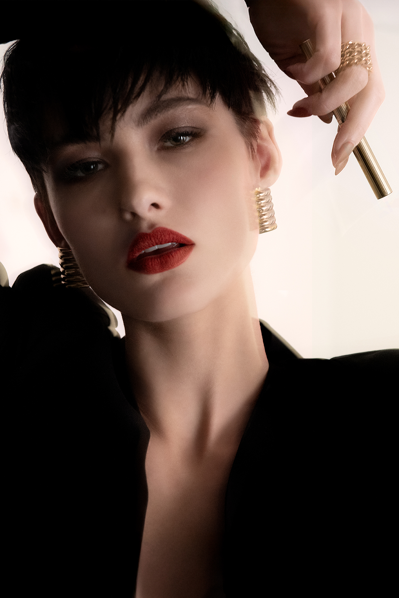 Close-up shot of a woman with short dark hair wearing gold earrings and a black blazer, holding a gold lipstick tube and ring.
