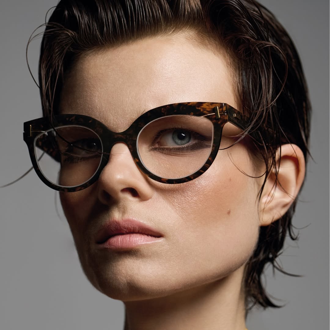 Close-up of a woman with fair skin and short, dark hair wearing dark-rimmed glasses, gazing into the camera with neutral expression.