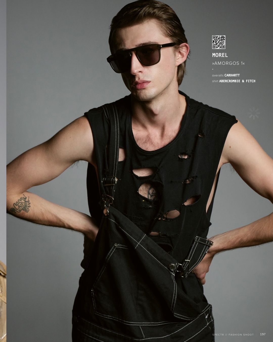 Fashion shot of a young adult male model posing in a distressed black tank top and dark overalls against a grey studio backdrop.