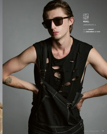 Fashion shot of a young adult male model posing in a distressed black tank top and dark overalls against a grey studio backdrop.