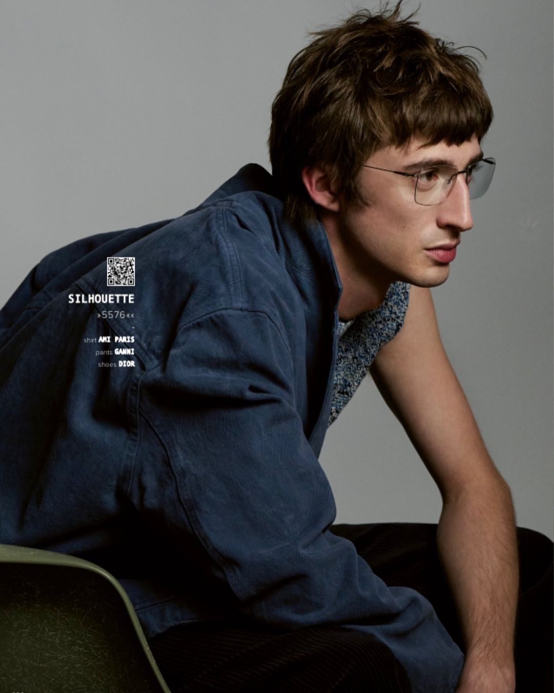 Portrait of a young adult male with brown hair wearing glasses and a blue jacket, seated in a green chair against a grey background.