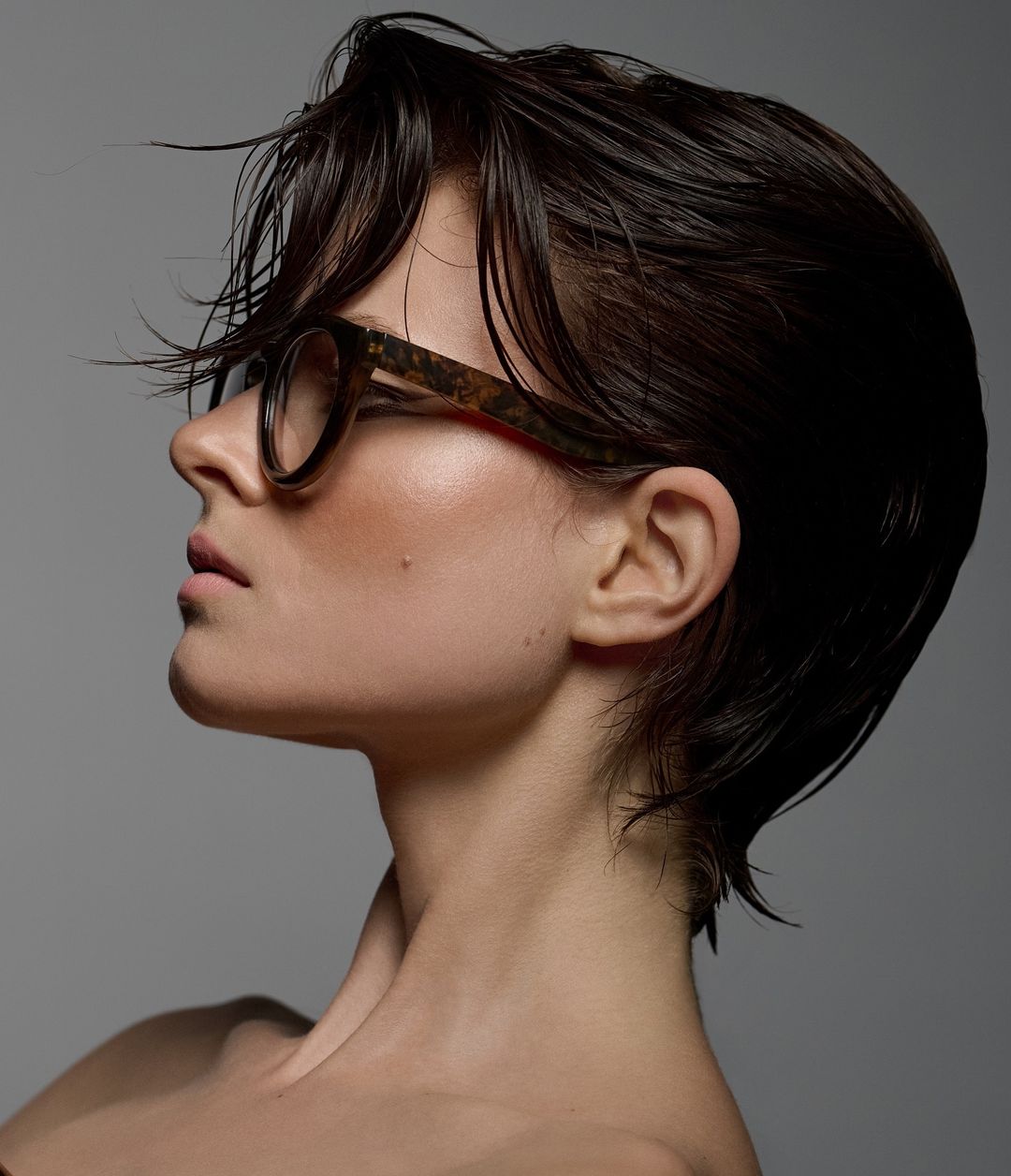 Headshot of a woman with wet dark hair and glasses in profile. She's facing left against a gray background in a beauty studio shot.