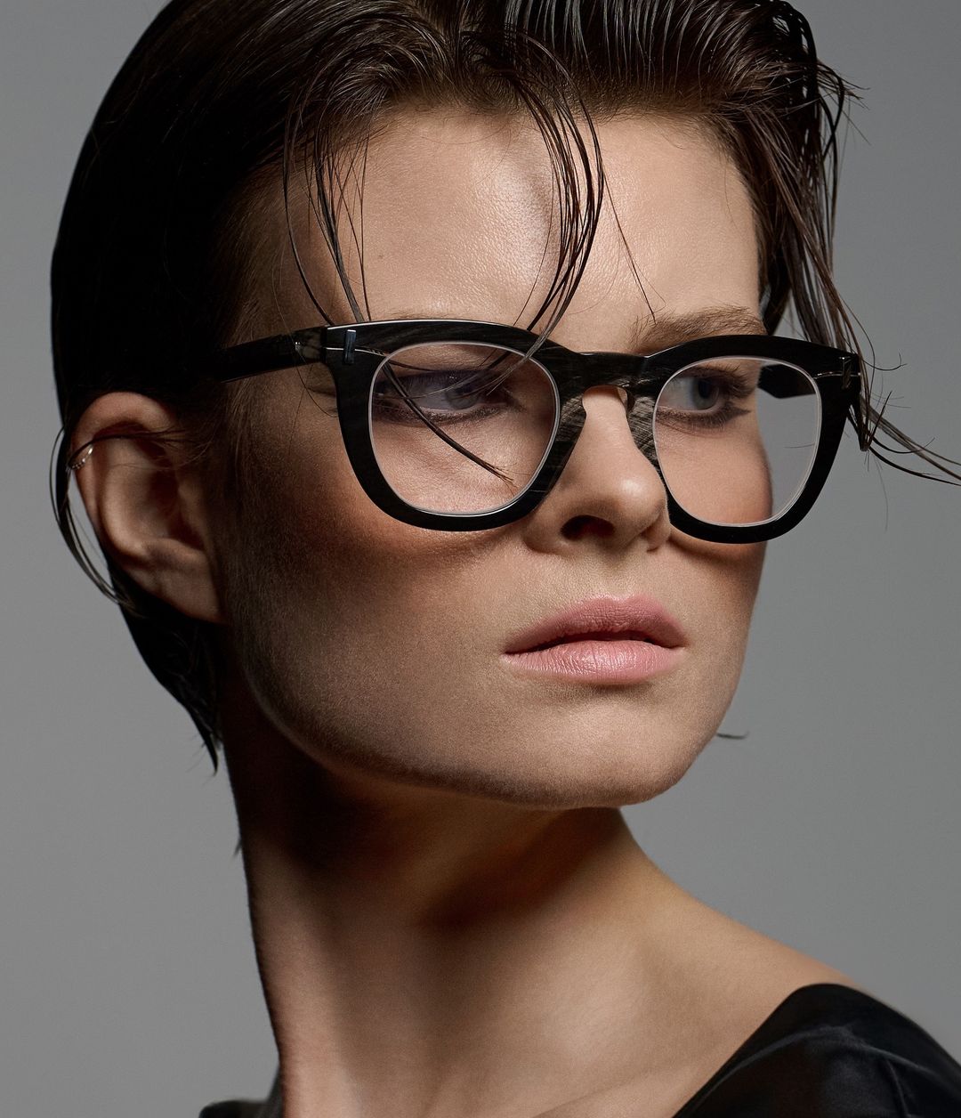 Close-up portrait of an adult female model with short, slicked-back hair, wearing black-rimmed glasses in front of a neutral gray backdrop.