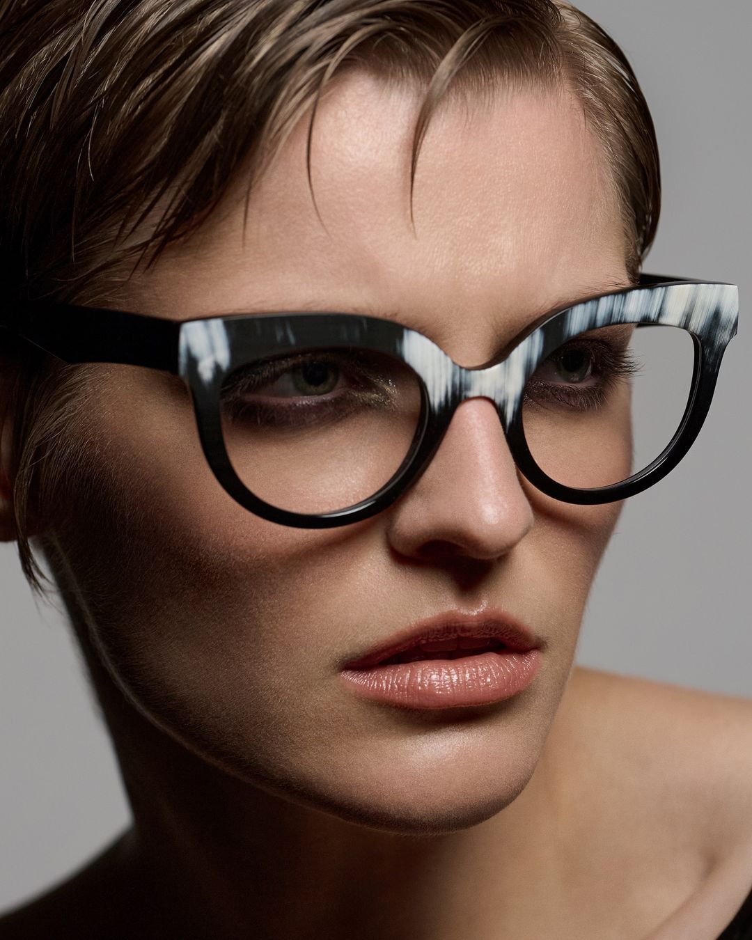 Close-up portrait of a fair-skinned woman with short hair wearing black and white cat-eye glasses against a grey backdrop.