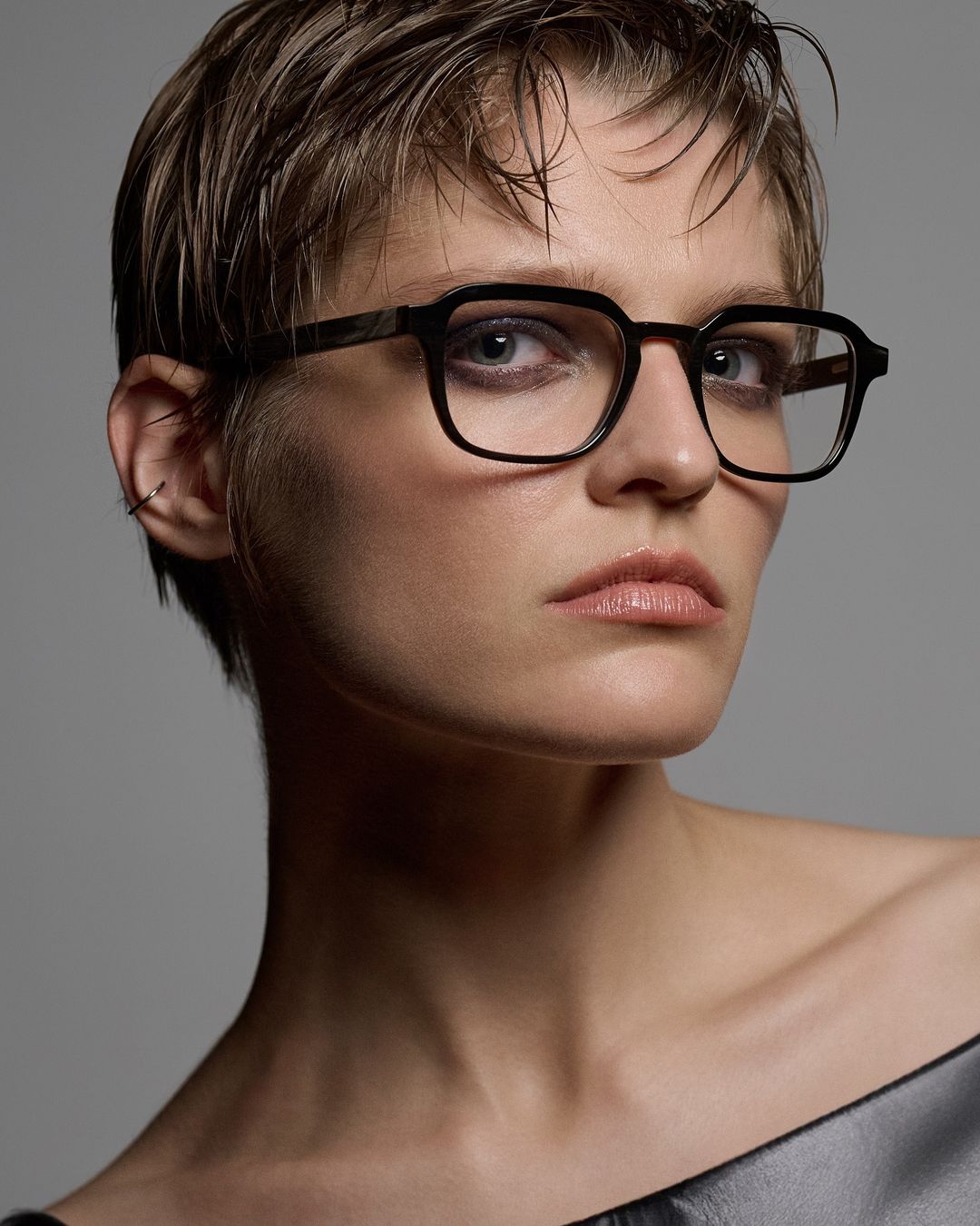 Close-up portrait of an adult woman with short blonde hair and wearing black-rimmed glasses in a studio with a gray backdrop.