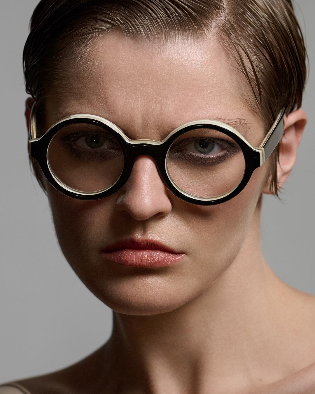 Close-up of an adult female wearing round eyeglasses with a black and white frame, posing in front of a neutral gray background.