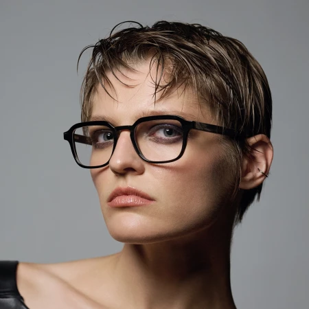 Close-up portrait of a woman with short wet hair and black framed eyeglasses against a plain grey background