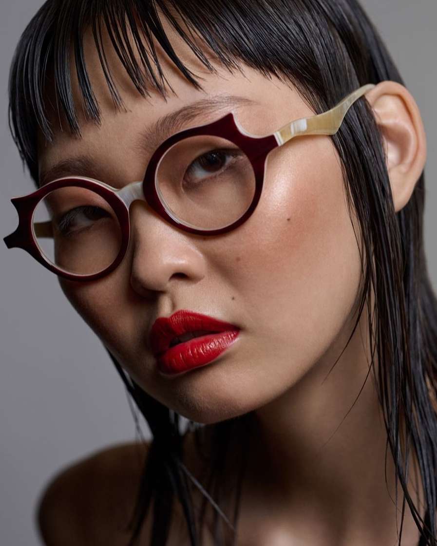 Close-up shot of a young Asian female model wearing round glasses with red frames and vibrant red lipstick against a gray background.