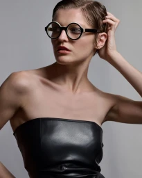 Model with short dark hair wearing round black and white glasses and a black strapless top against a grey background.