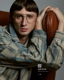 Man wearing wire-rimmed glasses and patterned shirt leaning against a brown leather chair