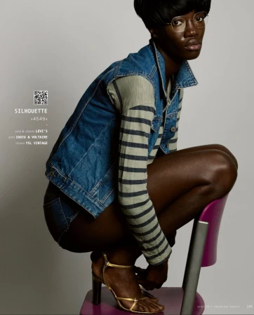 Black woman crouching on a purple chair wearing a denim vest, striped long-sleeve shirt, and gold heels against a grey background.