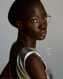 Black woman wearing thin wire-rimmed glasses and a metallic silver sleeveless top turning to look over her shoulder against a plain grey background