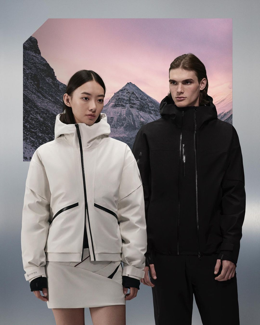 Two fashion models in ski jackets pose in front of mountain backdrop. Woman in white, man in black. Fashion shot. Studio lighting.
