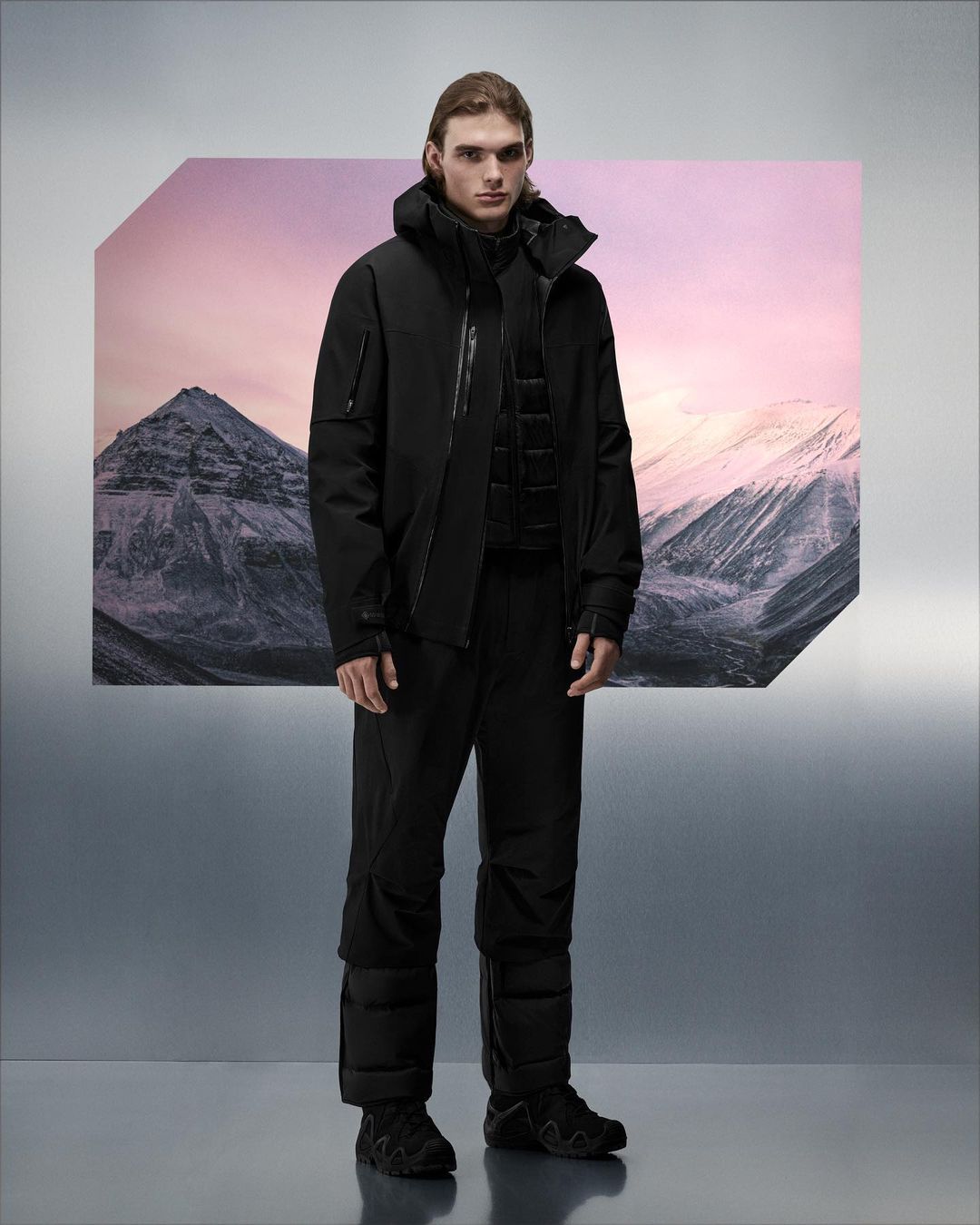 Full-length shot of a man posing in winter outerwear against a studio backdrop featuring snowy mountains and a pink sky.