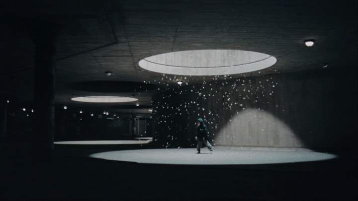 Child in hooded coat dances under beam of light in dark concrete interior with falling paper confetti creating dynamic scene.