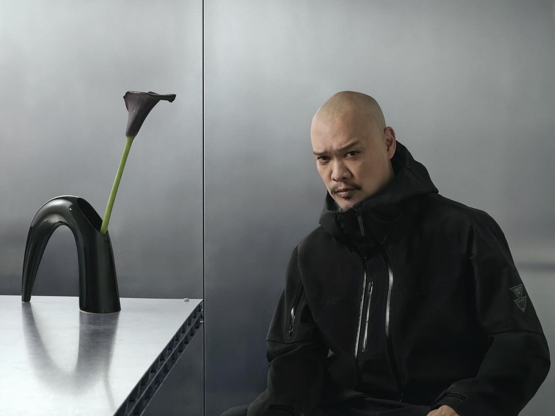 Portrait of a bald adult male in a dark hooded jacket, with a dark Calla lily in a black vase, and a silver metallic backdrop.