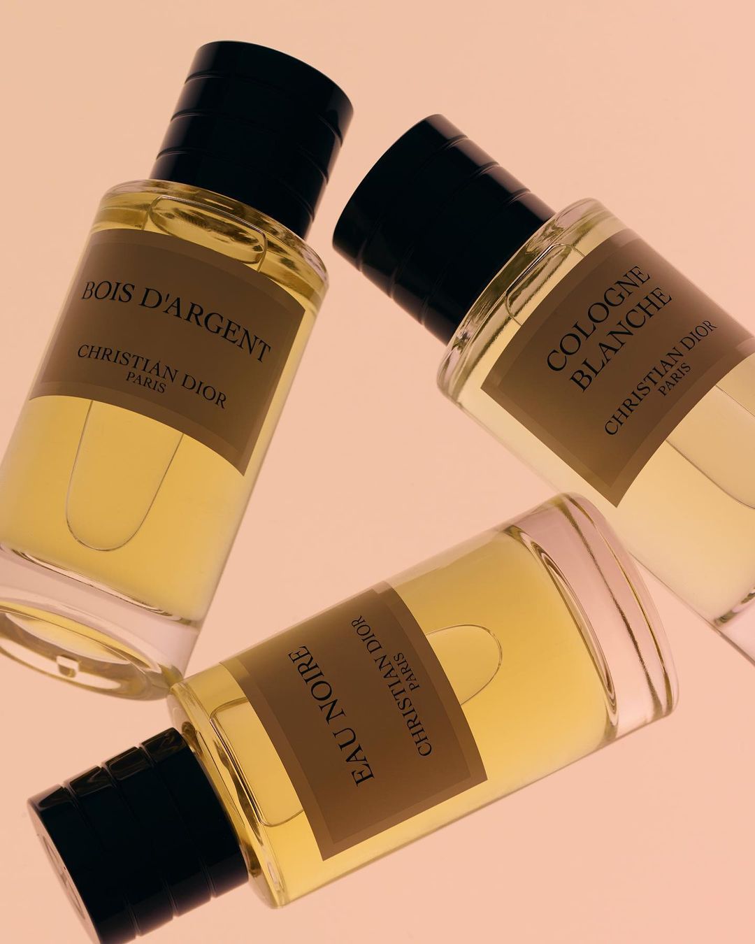 Three Christian Dior fragrances: Bois D'Argent, Eau Noire, and Cologne Blanche, displayed on a light pink surface in a beauty still life.