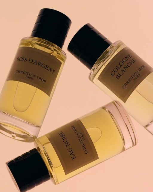 Three Christian Dior fragrances: Bois D'Argent, Eau Noire, and Cologne Blanche, displayed on a light pink surface in a beauty still life.