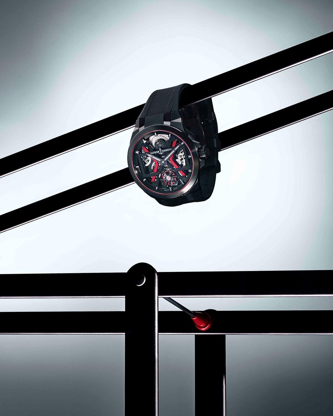 Luxury black Ulysse Nardin watch with red accents suspended by black rods on a white gradient background in a studio shot.