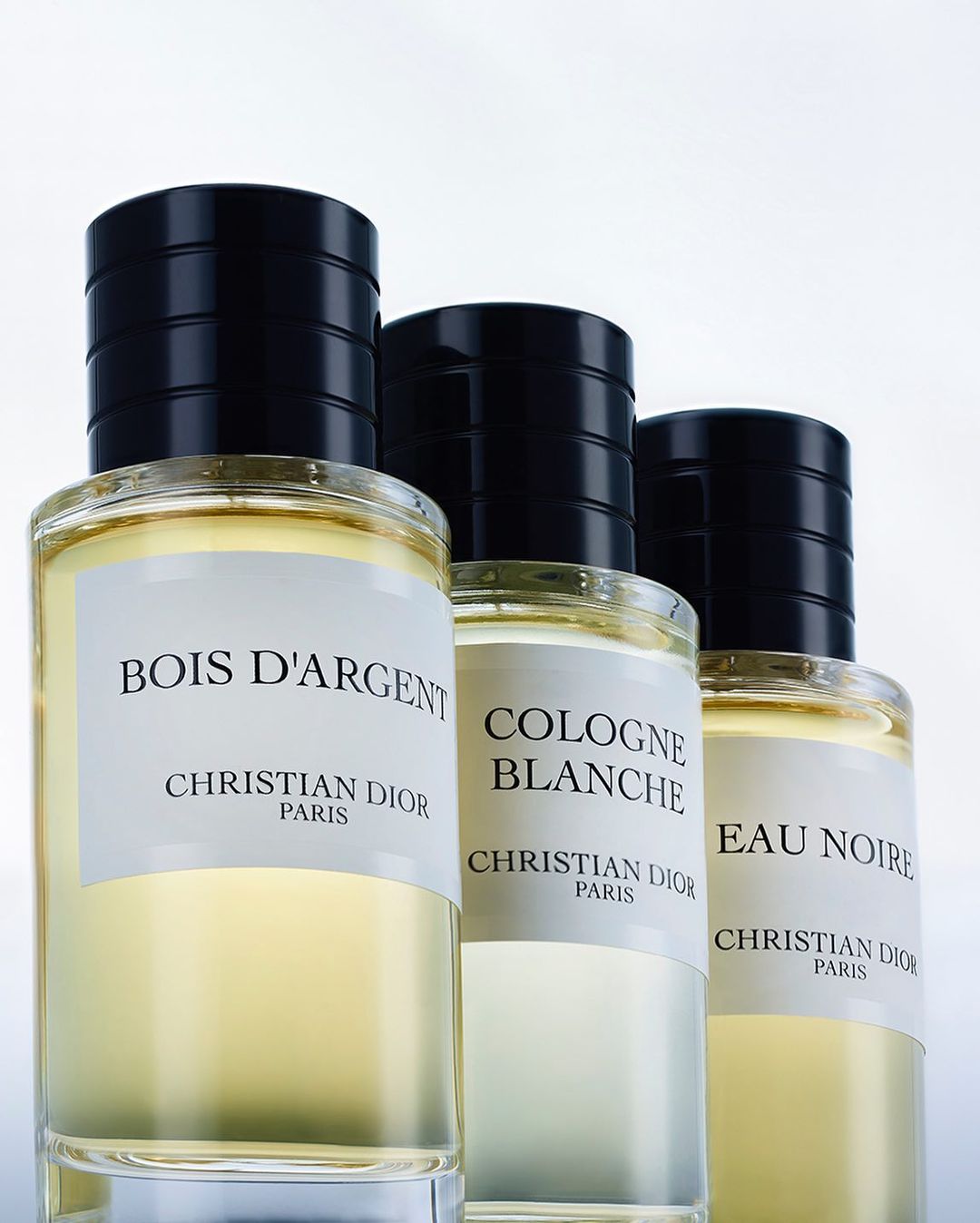 Three glass perfume bottles are lined up with labels facing forward, featuring black caps and clear liquid in a clean, brightly lit setting.