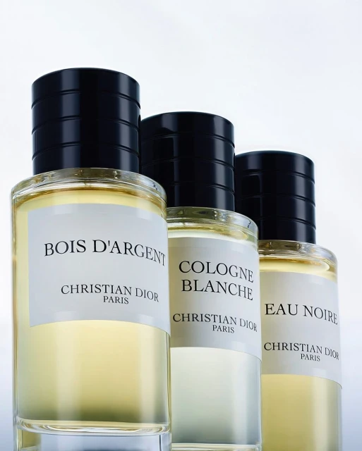 Three glass perfume bottles are lined up with labels facing forward, featuring black caps and clear liquid in a clean, brightly lit setting.
